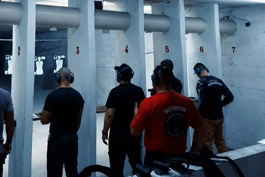 Instrutor de camiseta vermelha orienta alunos com protetores auriculares na linha de tiro do estande