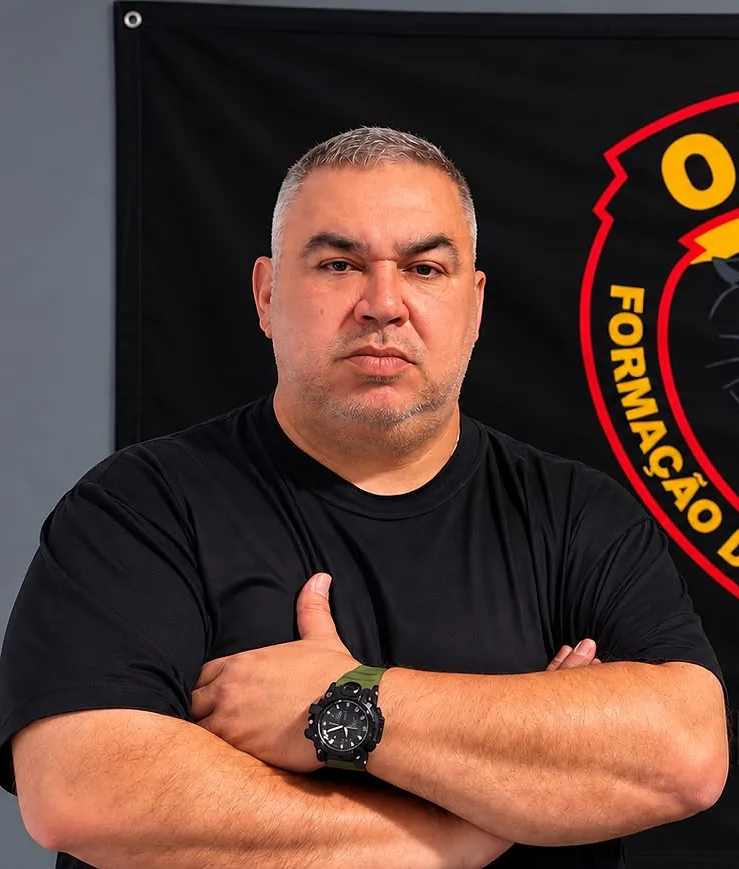 João Martins Arroyo Junior, sócio-fundador da OTMA, de braços cruzados diante da bandeira da escola