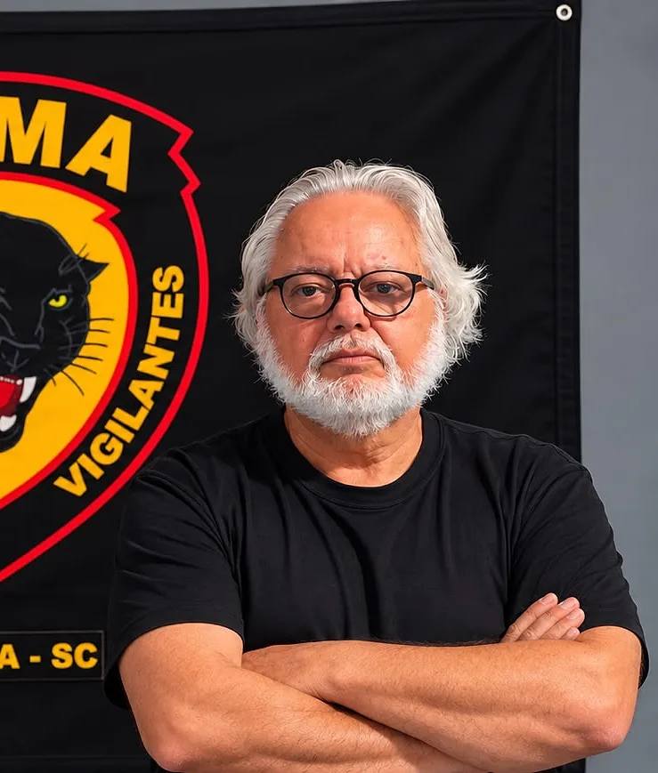 Othon da Silva Pias, sócio-fundador da OTMA, de braços cruzados diante da bandeira da escola