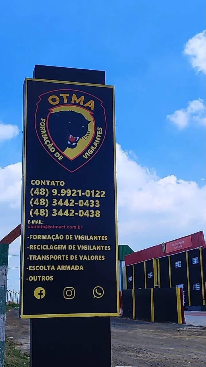 Placa da OTMA na entrada da escola com telefones de contato e os cursos de formação, reciclagem, transporte de valores e escolta armada